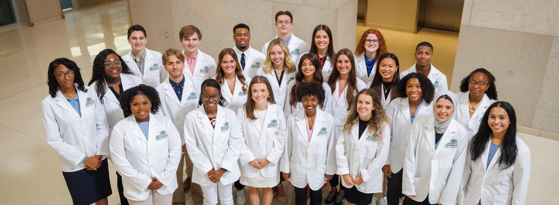 Large group of medical students in white lab coats gathered indoors for a group photo in an academic hallway.